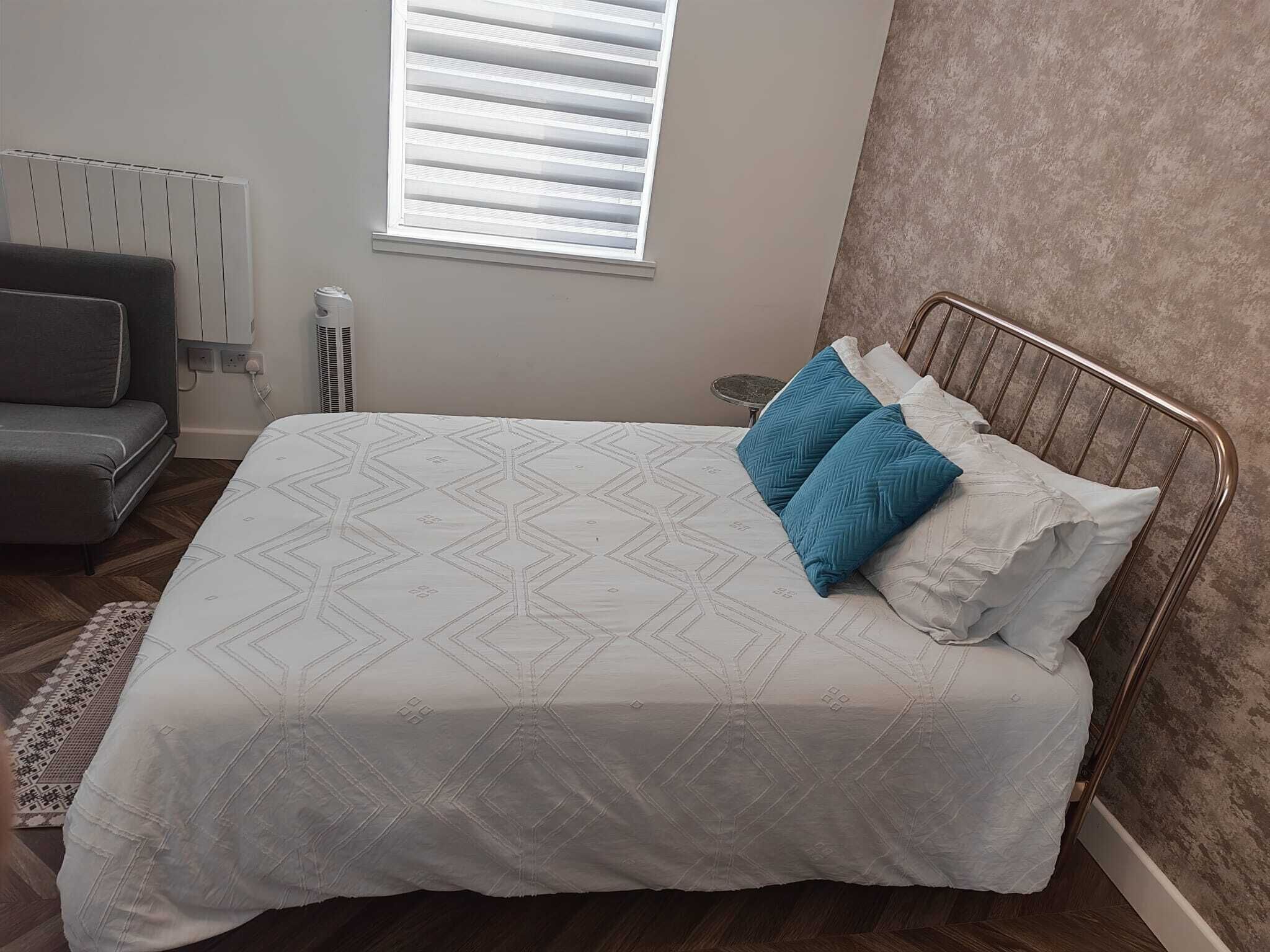 Une chambre à coucher avec un lit, un canapé gris, un radiateur et une fenêtre avec des stores.