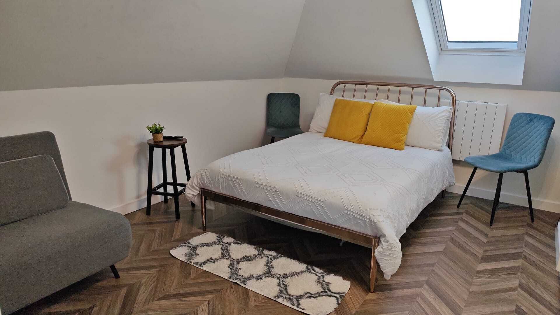 A bedroom with a bed, a grey sofa, a small table with a plant, a blue chair, and a patterned rug.
