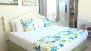 Un lit bien fait, recouvert d’une couvre-lit à motifs floraux, un ventilateur de plafond et un bureau avec une chaise, le tout dans une pièce dont le sol est carrelé.