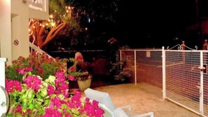 Un espace extérieur aménagé avec des chaises blanches et une table, entouré d’une clôture blanche et de plantes en pot ornées de fleurs roses.