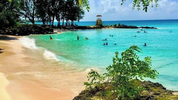A beach with clear turquoise water, a rocky foreground, and a lifeguard tower in the distance. - Villa Mia 2bed Apartment # 3 (Oistins)