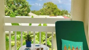 A balcony with a green cushioned chair, a small table with a potted plant, sunglasses, and a heart-shaped object. - Villa Mia 2bed Apartment # 3 (Oistins)