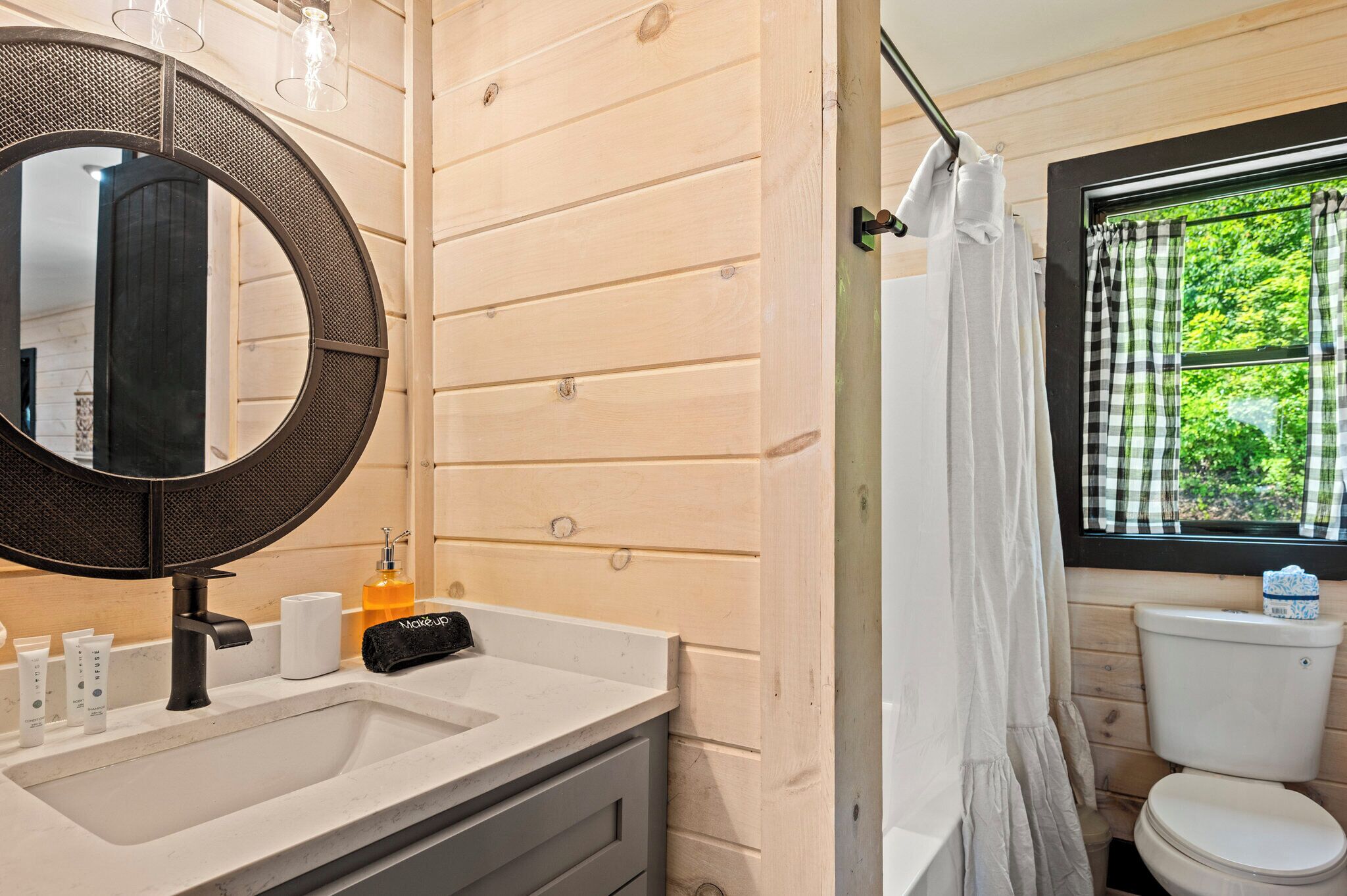 A bathroom with a wooden wall, a round mirror, a sink with toiletries, a toilet, and a window with a view of greenery.