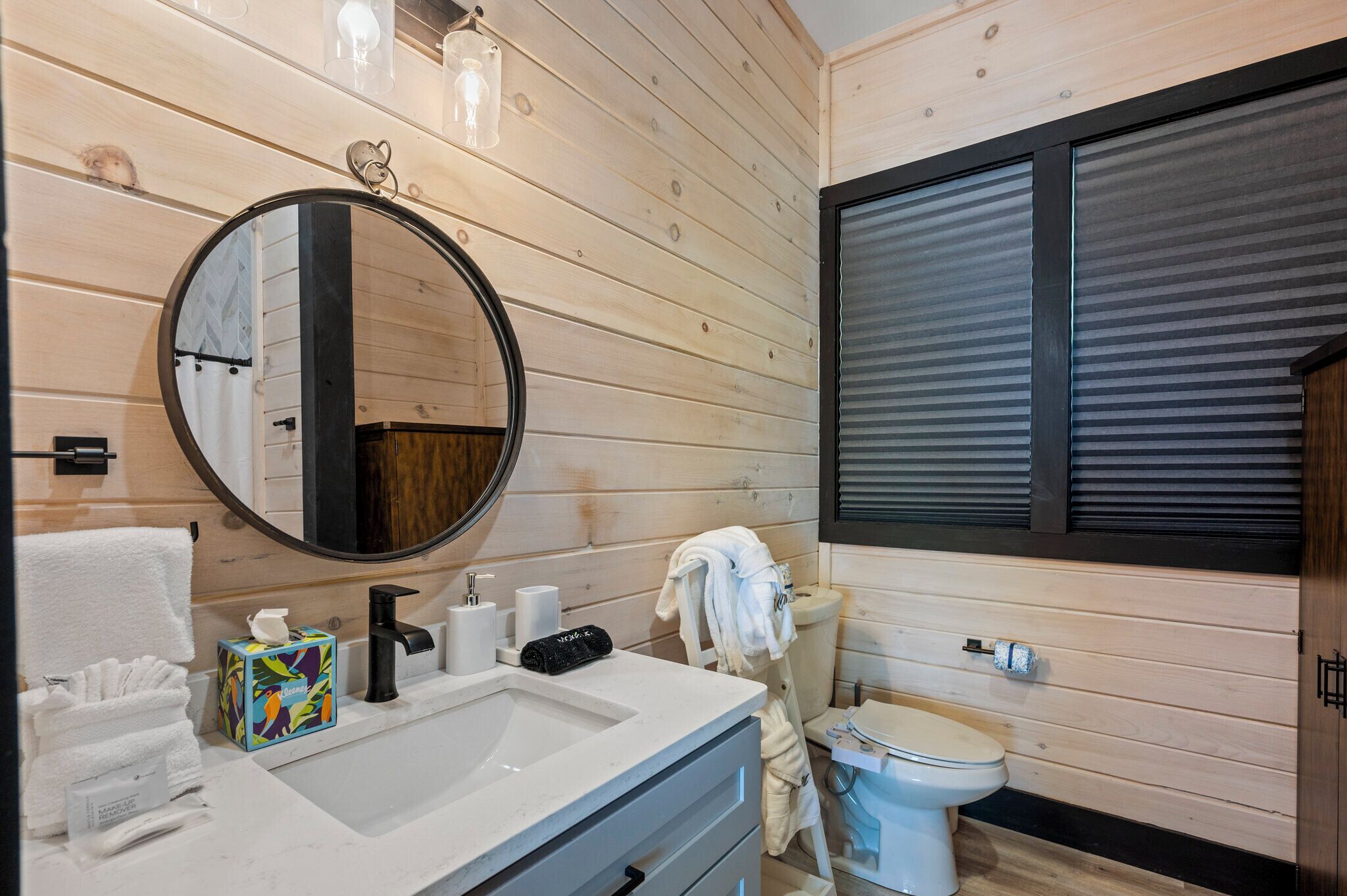 A modern bathroom with wooden paneling, a round mirror, a white sink, a toilet, and a window with blinds.