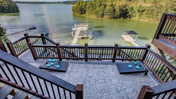 A deck with a view of a lake and dock, wooden railings, and a small table with chairs. - Stunning Mtn Lodge on Lake with two Story Dock, Indoor Pool, Arcade,and Hot Tub. (Sevierville)