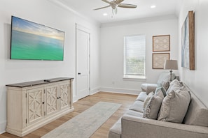 A modern living room with a large flat-screen TV, a ceiling fan, a wooden cabinet, a sofa with patterned cushions, and a window with blinds.