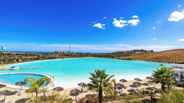 Un resort con una piscina grande, zona de playa con sombrillas y vistas al océano.