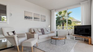 Una sala de estar moderna con un sofá, sillón, mesa de centro, televisor y ventanas amplias con vista a palmeras.