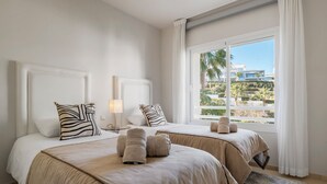 Habitación de hotel con dos camas, un ventanal con vistas a palmeras y un edificio, y una mesita con una lámpara.