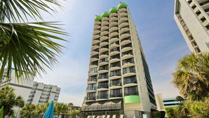 A tall, multi-story hotel with balconies, surrounded by palm trees and a pool area.