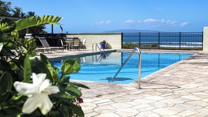 A swimming pool with a view of the ocean, surrounded by lounge chairs and a stone patio. - Ocean Front Condo, Modern & Bright (kihei)