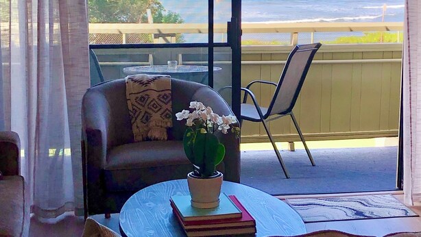 A room with a large sliding glass door leading to a balcony overlooking a beach. - Ocean Front Condo, Modern & Bright (kihei)