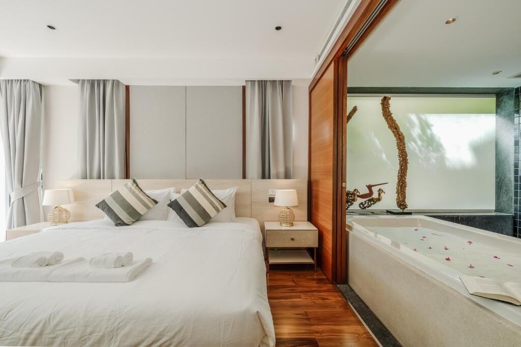 A modern hotel room with a large bed, a nightstand, a wooden floor, a large window, and a bathtub with rose petals.