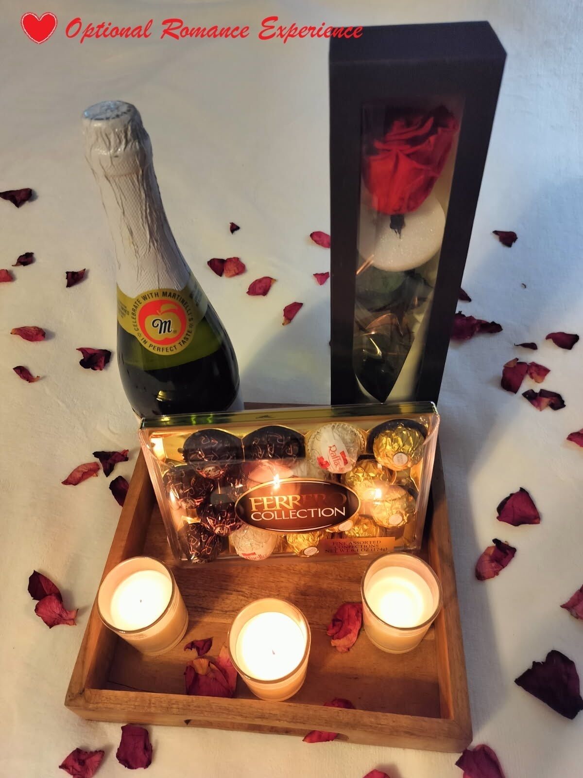 A bottle of champagne, a box of Ferrero Collection chocolates, and two lit candles on a wooden tray with rose petals scattered around.