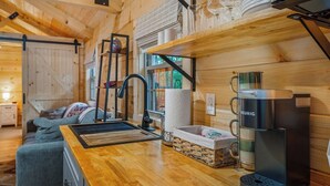 Un acogedor interior de cabaña con detalles de madera, una kitchenette con cafetera Keurig y un comedor con copas de vino.