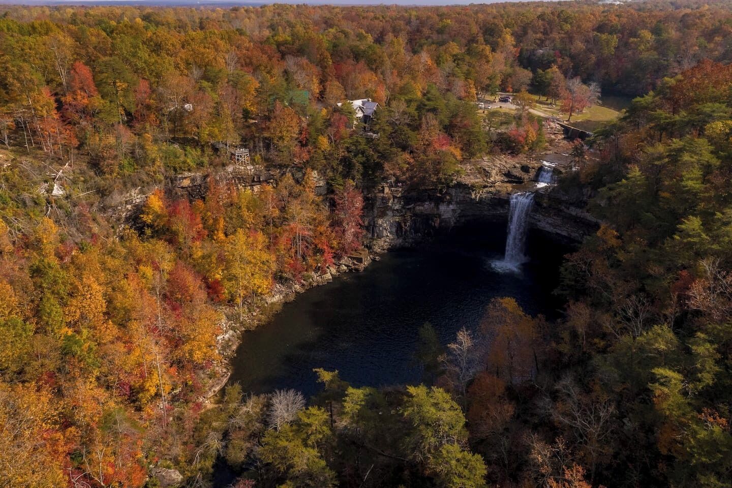 Ein Wasserfall, umgeben von Bäumen in Herbstfarben, mit einer rustikalen Holzhütte und einer Aussichtsplattform in der Nähe.