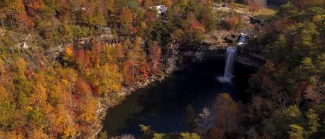 Ein Wasserfall, umgeben von Bäumen in Herbstfarben, mit einer rustikalen Holzhütte und einer Aussichtsplattform in der Nähe.