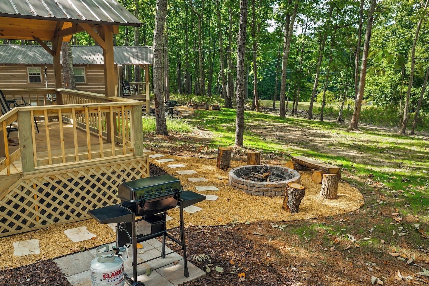 Une terrasse en bois avec un barbecue et une bouteille de propane, entourée d’un foyer et de bûches, avec une zone boisée en arrière-plan.