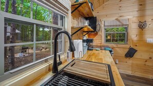 A wooden cabin interior with a kitchen area, including a sink, countertop, and a window overlooking a forest.