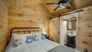 A wooden cabin bedroom with a bed, pillows, a ceiling fan, a nightstand, a mirror, and a bathroom with a sink and cabinet.