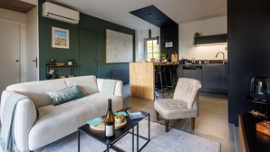 A modern living room with a sofa, armchair, and coffee table. There is a kitchen area with a dining table and chairs visible through an open door.