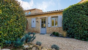 A stone house with a gravel driveway, surrounded by lush greenery and a variety of plants.