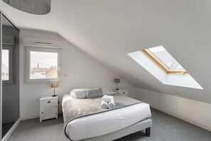 A modern bedroom with a skylight, a bed with white linens, a nightstand, and a view of the city through a window. - Cap Vauban - Duplex à 200m de la plage (Saint-Malo)