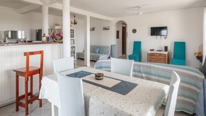 A modern kitchen with white cabinetry, a dining area with a striped tablecloth, and a living room with a sofa and teal chairs.