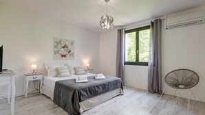 A bedroom with a bed, bedside tables, a hanging light fixture, a window with curtains, and an air conditioning unit.