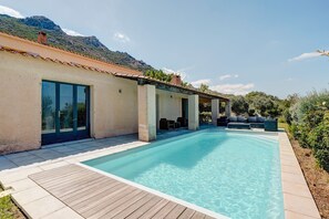 Un área junto a la piscina con una terraza de madera, mobiliario exterior y montañas al fondo.