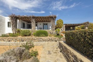 Een traditionele villa in mediterrane stijl, met een stenen pad dat naar de ingang leidt, omgeven door goed onderhouden hagen en verschillende struiken.