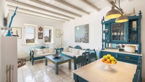 A cozy living room with a sofa, coffee table, and a dining area with a table and chairs. There is a window with a view of the outside, a painting on the wall, and a blue cabinet with kitchen items. - Isterni | 3BR House with Sea View & BBQ | Blue Coral 2 (Paros)