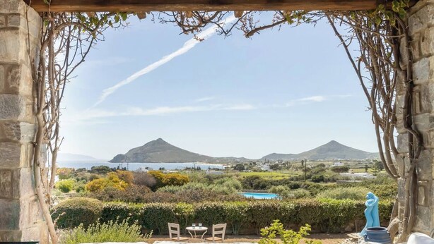 A covered outdoor seating area with a stone wall, a table with a fruit bowl, and a view of a mountainous landscape. - Isterni | 3BR House with Sea View & BBQ | Blue Coral 2 (Paros)