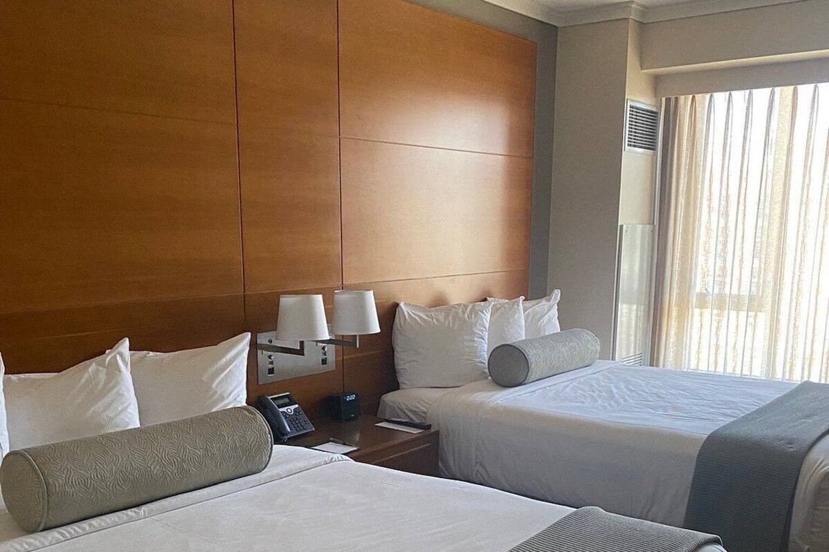 A hotel room with two beds, a wooden headboard, bedside lamps, and a window with curtains.