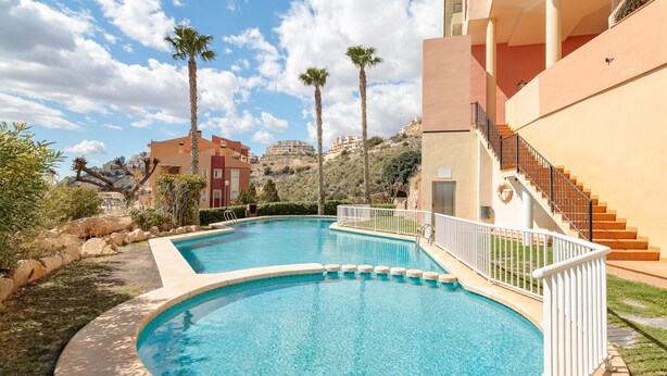 A swimming pool with a white railing, surrounded by greenery and palm trees, with buildings in the background. - Apartment 'El Mirador' with Sea View, Wi-Fi and Air Conditioning (Mutxamel)