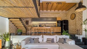 A modern living room with a white sectional sofa, a wooden coffee table, and a kitchen area with a brick wall and wooden beams.