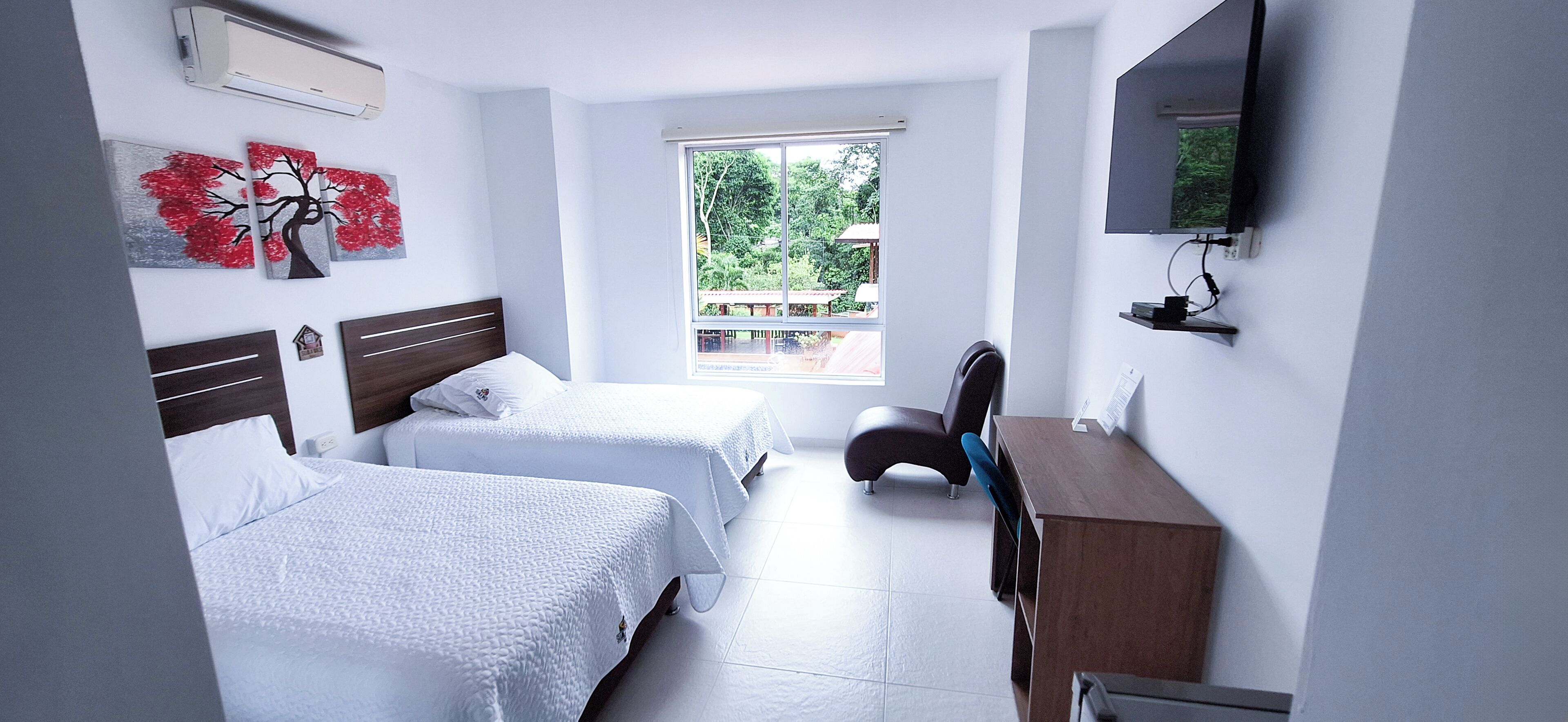 A hotel room with two beds, a desk, a chair, a TV, and a window with a view of greenery.