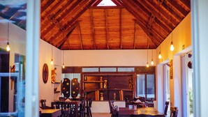 A restaurant with wooden ceiling beams, hanging light bulbs, and a central display shelf. Tables and chairs are arranged for dining. - HOTEL SUMAWASI (Mocoa)