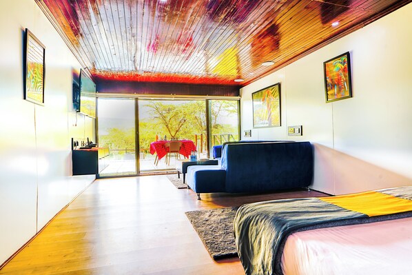 A modern bedroom with a large bed, a blue sofa, and a glass door leading to an outdoor area with a red table. - Kitumo Mara Lodges  (Maasai Mara)