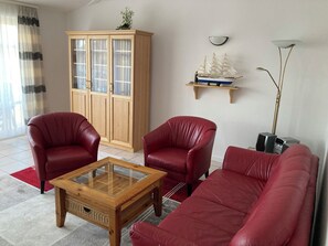 A living room with a wooden cabinet, two red leather chairs, a wooden coffee table, a red sofa, a ship model on a shelf, and a tall floor lamp. - 60sqm vacation apartment for 2 people in the Baltic seaside resort of Rerik (Rerik)