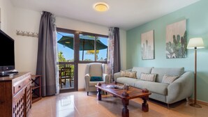 A living room with a sofa, armchair, coffee table, TV, and artwork on the wall. - Apartment 'Oasis Bahia Poolfront' with Shared Pool, Private Terrace & Wi-Fi (Corralejo)