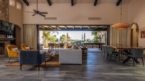 A spacious living room with a large TV, comfortable seating, a dining area, and a view of palm trees and a pool.