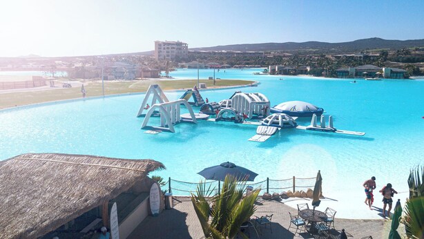 A large swimming pool with various water play structures, a thatched-roof building, and a view of the surrounding landscape.
