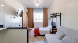 Una habitación de hotel pequeña con cama, sofá, televisor montado en la pared y ventana con cortinas.