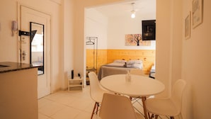 A compact hotel room with a kitchenette, a bed with a wooden headboard, a round dining table with chairs, and a small seating area.