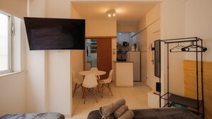 A modern living room with a flat-screen TV, a dining area with a small table and chairs, and a kitchenette in the background.