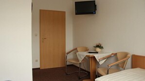A hotel room with a wooden door, a television mounted on the wall, two chairs, a small table with a vase of flowers, and a bed. - Gästehaus Zum Felsenkeller Steigerthal  (Nordhausen)