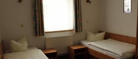 A small, simple room with two single beds, a chair, and a window with curtains.