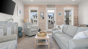 Un salón con un sofá gris, un sillón a rayas, una mesa de centro y un televisor montado en la pared. Hay una puerta de madera con un letrero encima y una ventana con persianas.