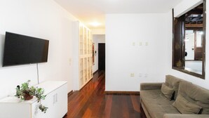 A room with a flat-screen TV mounted on the wall, a white cabinet with a plant, a beige sofa, and a wooden floor. - SUH Catete 06 Big House Metro (Rio de Janeiro)